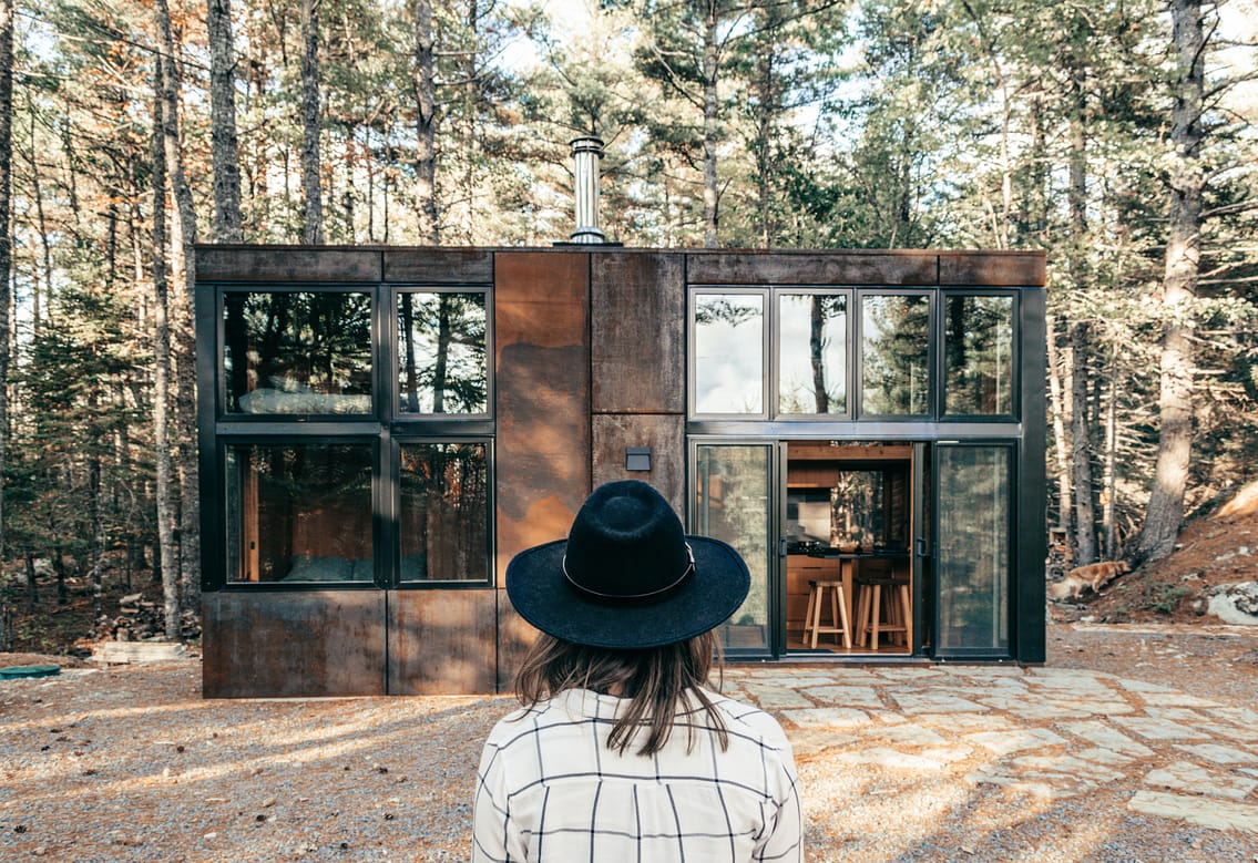 tiny house in the forest, woman with black hat sitting in front of the house