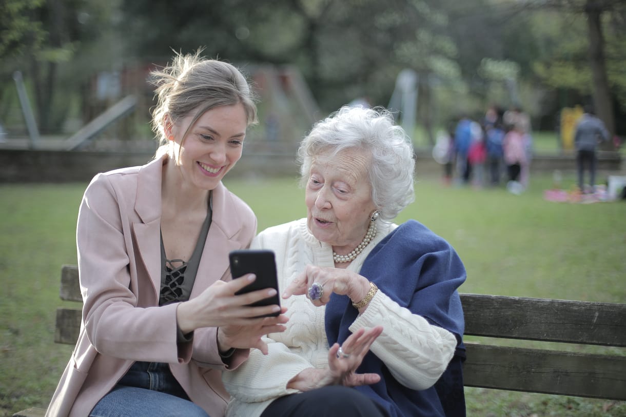 mature woman and younger woman, middle age sitting on a bench in the park, looking on a smartphone