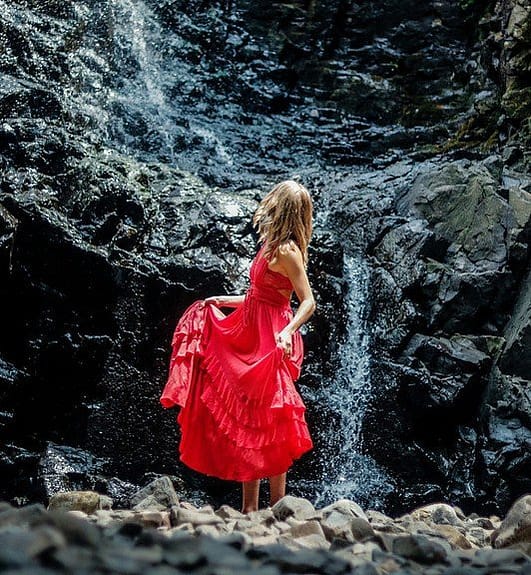 a woman dressed in a red dress in front of a small waterfall coming down a graphit grey wall